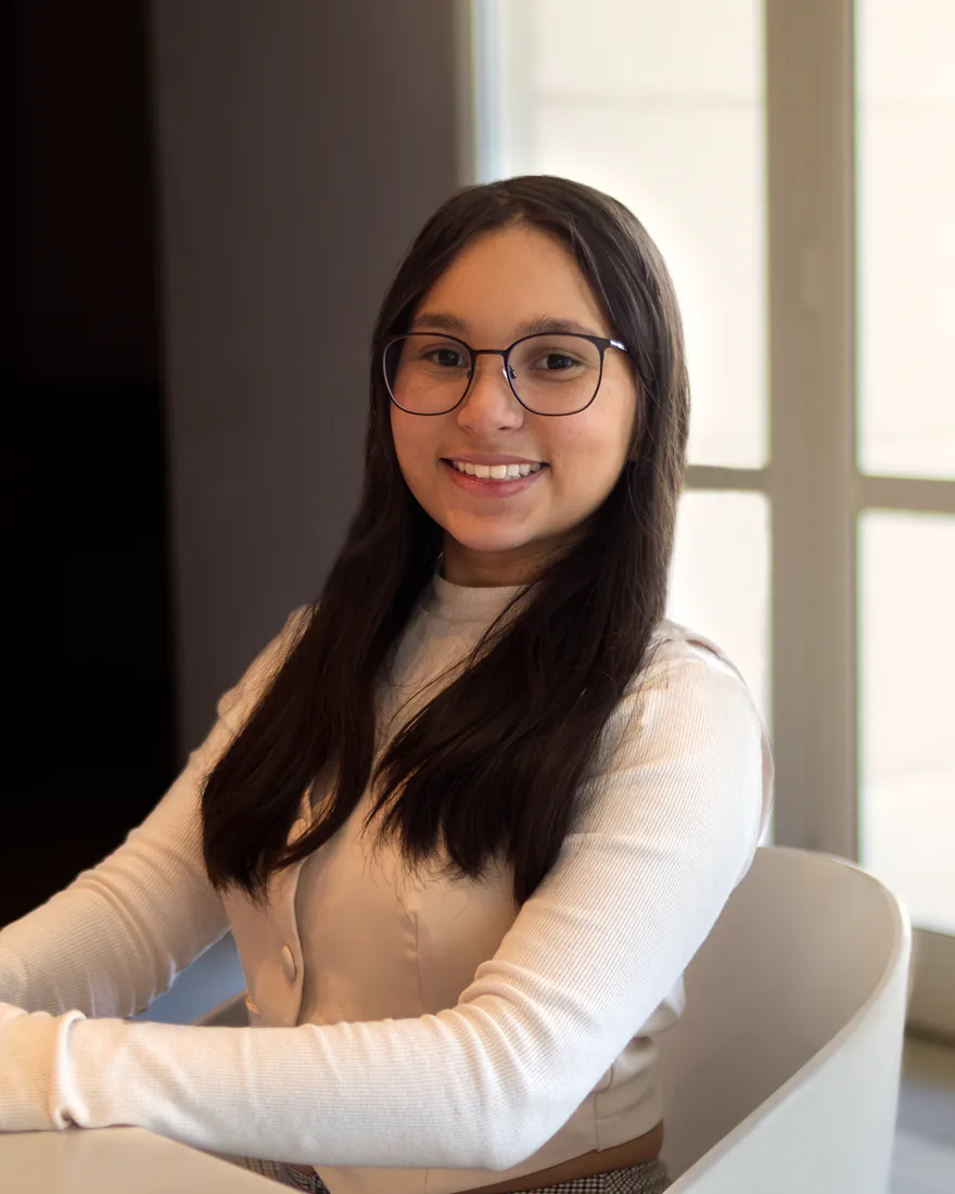 Maria Clara sorrindo para a câmera, sentada a uma mesa clara com uma janela ampla ao fundo.