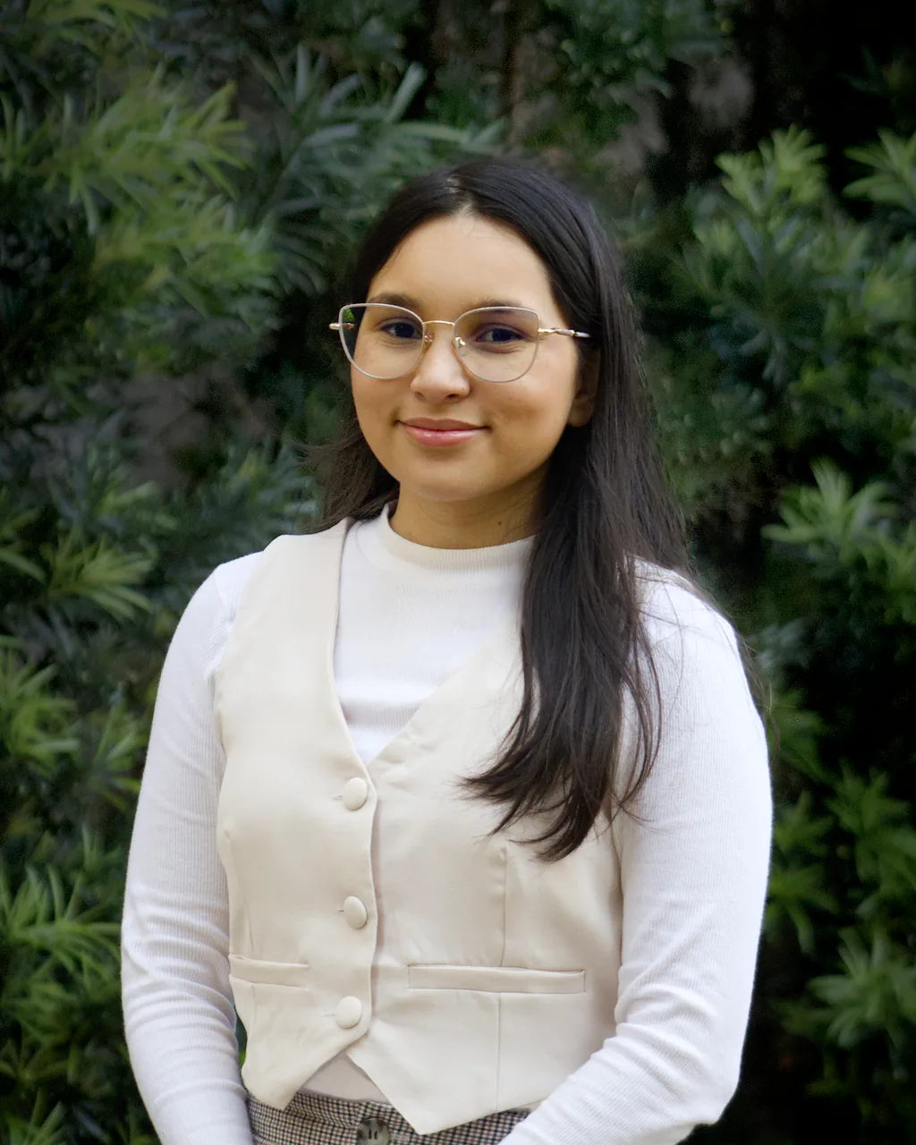 Maria Clara em pé ao ar livre, diante de uma cerca viva verde, de colete creme e sorriso leve.